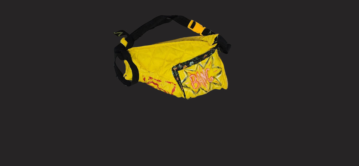 A yellow and black fanny pack with a graphic print on the front pocket, shown on a white sheet of paper next to a ruler and a person's foot.